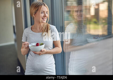Happy healthy pregnant young blond woman standing leaning against a patio window smiling as she looks outdoors while enjoying a bowl of fresh fruit sa Stock Photo