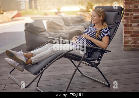 Happy smiling attractive young pregnant young woman relaxing outdoors on a recliner chair on a wooden deck cradling her baby bump in her hands Stock Photo