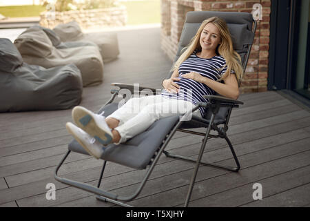 Happy smiling attractive young pregnant young woman relaxing outdoors on a recliner chair on a wooden deck cradling her baby bump in her hands Stock Photo