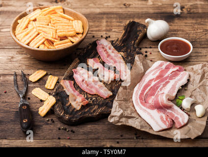 Bacon flavored snacks chips with grilled bacon rashers on vintage chopping board with smoked raw bacon on butchers paper with garlic and sauce on wood Stock Photo