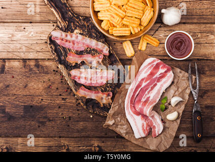 Bacon flavored snacks chips with grilled bacon rashers on vintage chopping board with smoked raw bacon on butchers paper garlic and sauce on wood back Stock Photo