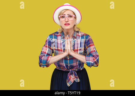 Please forgive me. or give me one more chance, Portrait of hopeful mature woman in casual style with hat and eyeglasses standing with palm hands and l Stock Photo