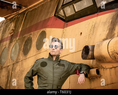 A handsome young man pilot in a green overall standing next to an old plane on a sunny day. Stock Photo