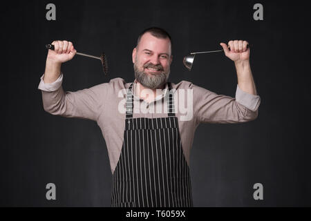 Mature european chef with beard holding a ladle standing on black studio wall. Stock Photo