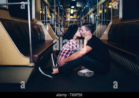 Young man and woman use underground. Couple in subway. Lovely cheerful couple sit together on floor and kissing. Alone in empty underground carriage.  Stock Photo