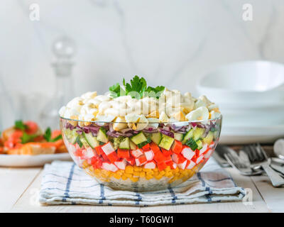 Large glass bowl with crab salad, corn, cucumber and rice at table. Layered crab sticks and corn salad. Traditiolnal mayonnaise salad for russian holiday feast, including new year. Stock Photo