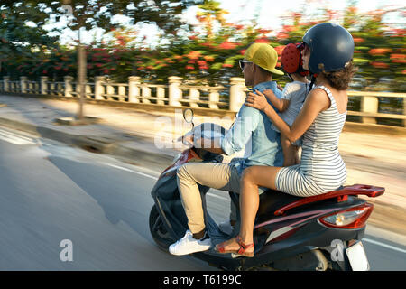 Cute couple with a small kid are riding red motorbike on the city street on sunny sky background in Vietnam. They wearing casual clothes, sunglasses,  Stock Photo