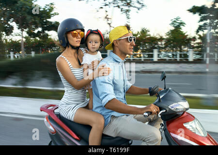 Glad couple with a small daughter are riding red motorbike on the city street on sunny sky background in Vietnam. They wearing casual clothes, sunglas Stock Photo