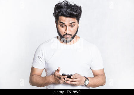 Perplexed and worried handsome young man holds smartphone, stares with surprised expression. Stock Photo