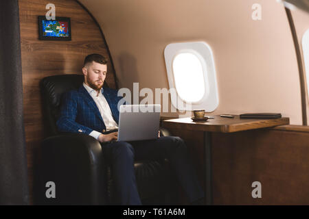 Attractive and successful businessman working on a laptop while sitting in a chair of his private jet. Stock Photo