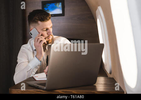 Young attractive and successful businessman talking on the phone and working while sitting in the chair of his private business plane Stock Photo