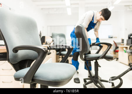 Young man in workwear and rubber gloves cleans the office chair with professional equipment. Stock Photo