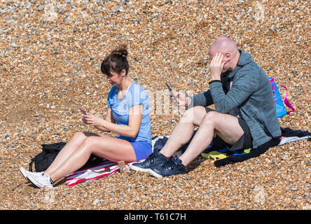 Couple of people in their 30's sitting on a beach on a warm day in Spring, ignoring each other while using mobile phones, in England, UK. Stock Photo
