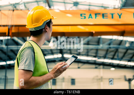 Young Asian male heavy industral worker using a digital tablet inside manufacturing and factory site with crane behind Stock Photo