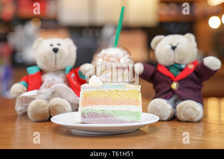 Slided rainbow cake decorate with unicorn horn on top next to couple teddy bear dolls. The cake is in a white disk on the wooden table and blur backgr Stock Photo