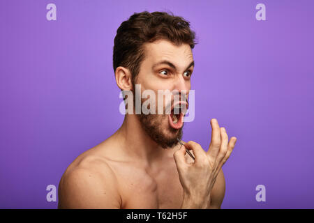 a suffering man plucking out his beard hair,standing isolated over blue background,close up side view photo. man is shouting as he feels pain while pl Stock Photo