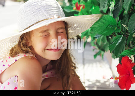 Beautiful little Girl looking at red flower and smiling in summer park. Happy cute kid playing outdoors. Stock Photo