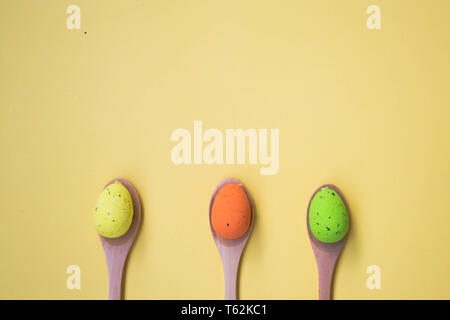 Three Colorful Easter eggs on pale yellow background, egg in wooden spoons, green, orange, yellow, egg hunt decoration, top view - flat lay Stock Photo