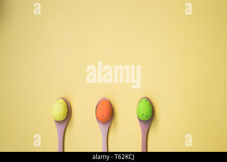 Three Colorful Easter eggs on pale yellow background, egg in wooden spoons, green, orange, yellow, egg hunt decoration, top view - flat lay Stock Photo