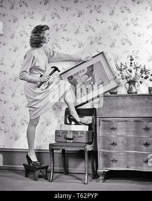 1940s DANGER WOMAN IN HIGH HEELS HOLDING HAMMER TRYING TO HANG PICTURE STEPPING CLIMBING FROM STOOL TO SUITCASE STACKED ON CHAIR - a3919 HAR001 HARS PERSONS DANGER RISK B&W STACKED STUPID TEMPTATION DISASTER WELLNESS DANGEROUS STRATEGY MISTAKE CHOICE IN ON TO CONCEPTUAL FRAMED HANG ATTEMPTING SOLUTIONS WRONG YOUNG ADULT WOMAN BLACK AND WHITE CAUCASIAN ETHNICITY HAR001 OLD FASHIONED Stock Photo