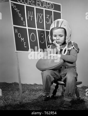 1960s DEJECTED BOY WEARING HELMET AND PADS SITTING HOLDING FOOTBALL IN FRONT OF HOME TEAM DEFEATED SCOREBOARD - b1098 HAR001 HARS OLD TIME NOSTALGIA OLD FASHION 1 JUVENILE FACIAL STYLE LOST COMPETITION SCORE ATHLETE LIFESTYLE FULL-LENGTH MALES ATHLETIC EXPRESSIONS B&W SADNESS PROTECTIVE SCOREBOARD DISASTER AND LEADERSHIP PADS PRIDE CONCEPTUAL IN FRONT OF LOSING BEAT DEJECTED EMOTION EMOTIONAL EMOTIONS GROWTH JUVENILES MISERABLE SEASON BLACK AND WHITE CAUCASIAN ETHNICITY DEFEATED HAR001 OLD FASHIONED Stock Photo