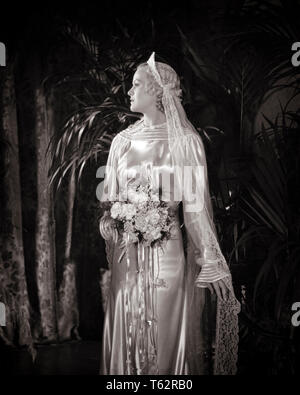 1930s BLONDE BRIDE IN PROFILE WEARING SATIN GOWN DRESS RIBBON RUFFLE NECK AND CUFFS HEADPIECE WITH VEIL FLORAL BOUQUET - b14896 HAR001 HARS EVENT COPY SPACE HALF-LENGTH LADIES MARRIAGE PERSONS INSPIRATION SATIN VEIL CONFIDENCE CEREMONY B&W BRIDAL BRIDES STYLES CUSTOM AND TRADITION CUFFS NUPTIAL NUPTIALS OCCASION MARRYING PRIDE HEADPIECE RITE OF PASSAGE RUFFLE STYLISH WED FASHIONS MARRY MATRIMONY YOUNG ADULT WOMAN BLACK AND WHITE CAUCASIAN ETHNICITY HAR001 OLD FASHIONED Stock Photo