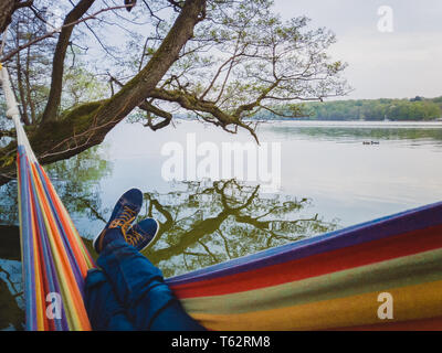 Man with blue shoes and jeans swings in colorful hammock hangs on shore of the lake in Denmark Stock Photo