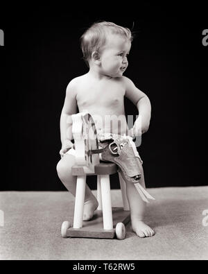 1930s 1940s BABY BOY SITTING ON TOY HORSE AND WEARING TOY WESTERN COWBOY GUN IN HOLSTER - b6592 HAR001 HARS VISION AND PRIDE IMAGINATION BABY BOY GROWTH JUVENILES BLACK AND WHITE CAUCASIAN ETHNICITY HAR001 LOOKING TO SIDE OLD FASHIONED Stock Photo