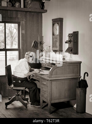 1920s 1930s SENIOR MAN WEARING VISOR SITTING IN OFFICE AT ANTIQUE WOODEN ROLLTOP DESK WITH WOODEN CRANK TELEPHONE ON WALL - bo00106 CAM001 HARS LIFESTYLE CLERK ELDER VERTICAL JOBS COPY SPACE HALF-LENGTH PERSONS MALES SENIOR MAN SENIOR ADULT B&W SKILL OCCUPATION SKILLS OLDSTERS OLDSTER OAK CAM001 OCCUPATIONS ELDERS KEROSENE ELDERLY MAN SALESMEN UMBRELLA STAND VISOR BLACK AND WHITE CAUCASIAN ETHNICITY OLD FASHIONED Stock Photo