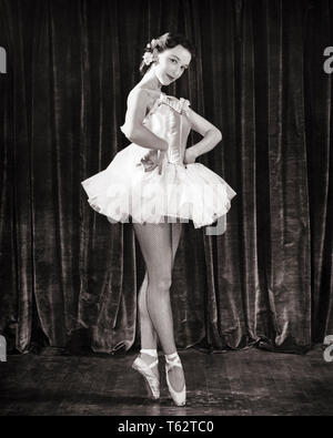 1950s BRUNETTE BALLERINA WEARING TUTU STANDING EN POINTE BEFORE STAGE CURTAIN LOOKING AT CAMERA HANDS ON HIPS - d2445 HAR001 HARS JOY LIFESTYLE FEMALES JOBS HEALTHINESS COPY SPACE FULL-LENGTH LADIES PERSONS ENTERTAINMENT CONFIDENCE B&W EYE CONTACT BRUNETTE PERFORMING ARTS SKILL BEFORE HIPS OCCUPATION HAPPINESS SKILLS STRENGTH TUTU PRIDE BALLERINA FEMININE OCCUPATIONS POSING STYLISH FISHNET STOCKINGS CREATIVITY EN POSED PRIMA TALENT YOUNG ADULT WOMAN BLACK AND WHITE CAUCASIAN ETHNICITY HAR001 OLD FASHIONED ON POINTE POINTE Stock Photo