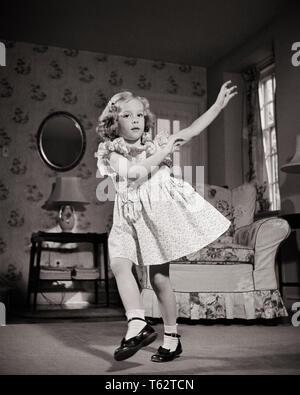 1940s BLOND GIRLIN DRESS WEARING MARY JANE SHOES LOOKING AT CAMERA DANCING HOKEY-POKEY IN LIVING ROOM  - d686 HBB001 HARS HOME LIFE UNITED STATES COPY SPACE FULL-LENGTH IRELAND B&W EYE CONTACT STRUCTURE AUSTRALIA PERFORMER LIVING ROOM EXCITEMENT RECREATION SONG MARY JANE PATENT LEATHER ENTERTAINER DISTINCTIVE BRITISH ISLES STYLISH MUSIC HALL ENTERTAINERS HOKEY POKEY JUVENILES PARTICIPATION PERFORMERS TUNE BLACK AND WHITE CAUCASIAN ETHNICITY LYRIC NOVELTY OLD FASHIONED Stock Photo