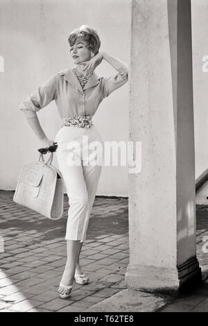 1950s 1960s FULL LENGTH PORTRAIT WOMAN WEARING BUTTON DOWN SHIRT CAPRI PANTS HOLDING STRAW BAG SUNGLASSES POSING BY COLUMN  - f6552 DEB001 HARS CHIN B&W STYLES SOPHISTICATED COLUMN BLOUSE OCCUPATIONS POSING COSMOPOLITAN STYLISH DEB001 DETACHED FASHION MODEL UNCONCERNED CAPRI CHIC FASHIONS MID-ADULT MID-ADULT WOMAN UNEMOTIONAL ATTITUDE BLACK AND WHITE BLASE CAUCASIAN ETHNICITY INDIFFERENT OLD FASHIONED Stock Photo