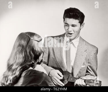 1950s COUPLE MAN WEARING SUIT AND TIE SINGING PLAYING UKULELE FOR WOMAN SEEN FROM BEHIND - m1825 CLE003 HARS LADIES PERSONS INSPIRATION MALES ENTERTAINMENT B&W SUIT AND TIE HAPPINESS HIGH ANGLE AND HAIRSTYLE SONG VOCAL REAR VIEW ENTERTAINER MUSICAL INSTRUMENT CONNECTION VOCALIZE CONCEPTUAL VOCALS LONG HAIR SONGS STYLISH UKULELE BACK VIEW CREATIVITY TOGETHERNESS YOUNG ADULT MAN YOUNG ADULT WOMAN BLACK AND WHITE CAUCASIAN ETHNICITY OLD FASHIONED Stock Photo