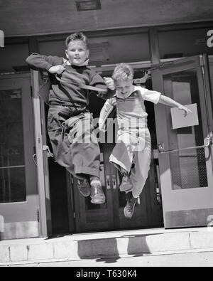 1950s two boys running jumping out the doors of ELEMENTARY school  - s617 HAR001 HARS HEALTHINESS COPY SPACE FRIENDSHIP FULL-LENGTH INSPIRATION MALES RELEASED SIBLINGS CONFIDENCE B&W FREEDOM SCHOOLS GRADE HAPPINESS CHEERFUL ADVENTURE LEAP EXCITEMENT OF THE FREE PRIMARY SIBLING SMILES ESCAPE FRIENDLY JOYFUL K-12 LIBERATED PANORAMIC GRADE SCHOOL GROWTH JUVENILES SCHOOLMATES TOGETHERNESS BLACK AND WHITE CAUCASIAN ETHNICITY CLASSMATES HAR001 OLD FASHIONED Stock Photo