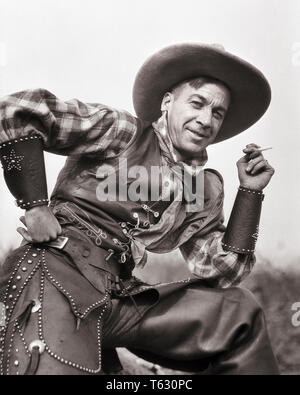 1920s AMERICAN COWBOY LEATHER DUDS VEST CHAPS WRIST CUFFS SMOKING A CIGARETTE LOOKING AT CAMERA - t6977 HAR001 HARS LIFESTYLE HISTORY JOBS RURAL HOME LIFE COPY SPACE HALF-LENGTH PERSONS MALES RISK WESTERN PROFESSION CONFIDENCE AMERICANA VEST EXPRESSIONS B&W EYE CONTACT CIGARETTES COWBOYS SKILL OCCUPATION HAPPINESS SKILLS CHEERFUL ADVENTURE CAREERS LOW ANGLE CUFFS PRIDE OCCUPATIONS SMILES JOYFUL STYLISH HAND ON HIP PLAID SHIRT CHAPS MID-ADULT MID-ADULT MAN BLACK AND WHITE CAUCASIAN ETHNICITY HAR001 OLD FASHIONED Stock Photo