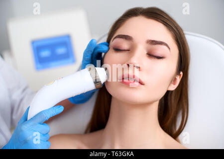Beautician doctor doing rf-lifting procedure for flawless woman face laying on medical chair in a beauty salon. Hardware cosmetology. Patient receivin Stock Photo