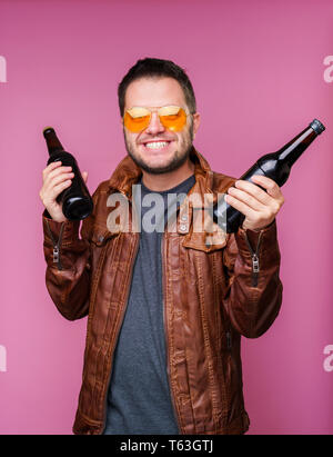 Photo of smiling man in orange glasses and leather jacket with two bottles in hands on empty pink background Stock Photo