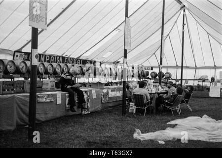 A 1970s photograph taken in England, showing a Beer Festival inside a canvas tent, run by CAMRA, the Campaign For Real Ale. Lots of barrels and casks of real ale beer seen stacked for serving with a table of men sitting around drinking and talking. Stock Photo