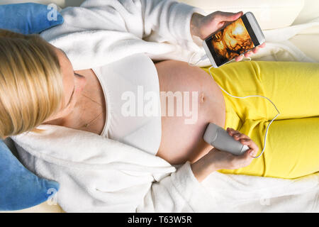 Modern pregnant woman sitting on a white couch at home relaxing and doing by herself digital 3D ultrasound scan of a baby with a mobile smart phone Stock Photo