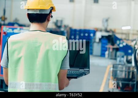 Asian male Industrial engineer in hardhat works with laptop in safety jacket at heavy industry factory. Processing plastic injection molding industry Stock Photo