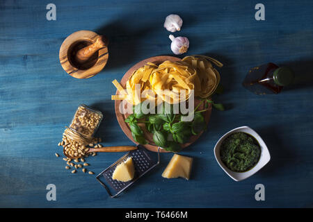 The table with ingredients to produce basil pesto. Mortar, fresh basil herbs, pine nuts, olive oil, parmesan cheese, garlic, bowl with pesto, grater,  Stock Photo