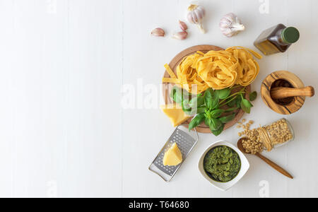 The table with ingredients to produce basil pesto. Mortar, fresh basil herbs, pine nuts, olive oil, parmesan cheese, garlic, bowl with pesto, grater,  Stock Photo