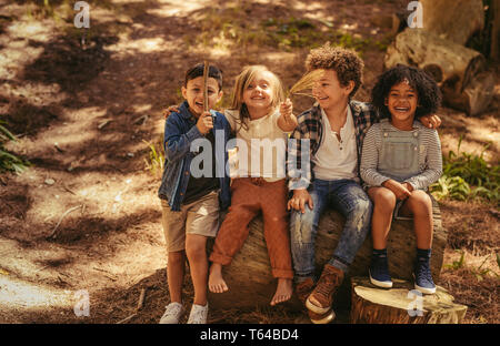 Group of boys and girl sitting on a log.  Cute kids outdoor enjoying nature. Children smiling and showing stick and twig. Stock Photo