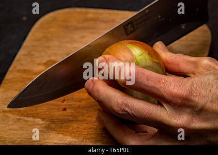 Close up of a chef chopping an organic brown onion (allium) with a sharp knife on a wooden chopping board. Stock Photo