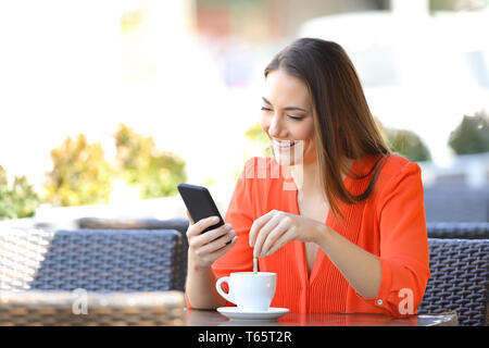 Happy woman using smart phone stirring coffee in a bar terrace Stock Photo
