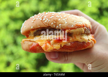 Melting cheese hamburger being held by hand against vibrant green foliage Stock Photo