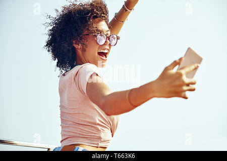 Carefree young woman wearing sunglasses and laughing while standing on a boat against a blue sky taking selfies during summer vacation Stock Photo