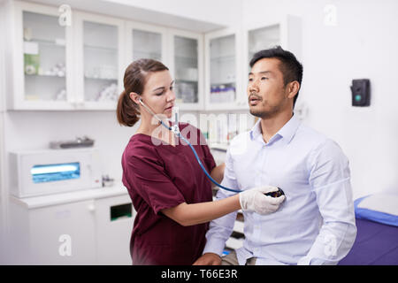 Female nurse using stethoscope on male patient in clinic examination room Stock Photo