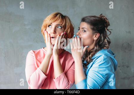 Two beautiful funny young women real friends in casual dress tell each other terrible secrets on concrete wall background Stock Photo