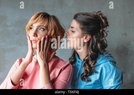 Two beautiful funny young women real friends in casual dress tell each other terrible secrets on concrete wall background Stock Photo